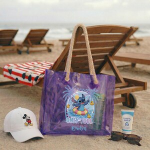 May include: A purple transparent tote bag with rope handles, featuring a Stitch graphic and the text "ONLY POOL Evelyn". A white baseball cap with a Mickey Mouse graphic, sunglasses, and sunscreen are nearby. Beach chairs and the ocean are in the background.