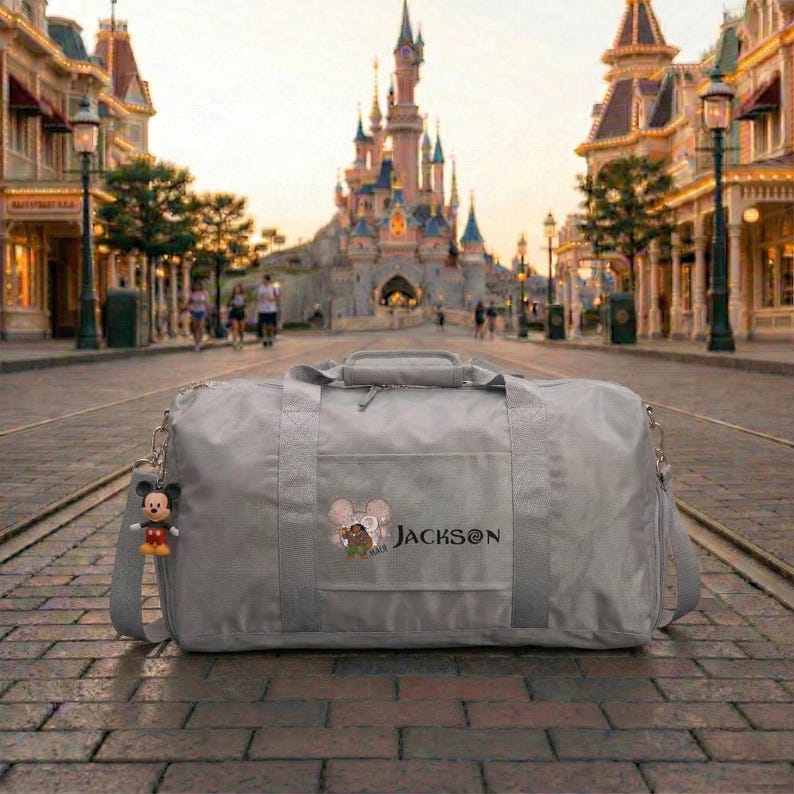 May include: A gray duffel bag with a Mickey Mouse keychain. The bag has a shoulder strap and a top handle. The bag has an embroidered design of Mickey and Minnie Mouse with the name "JACKSON" below. The background shows a castle and a street.