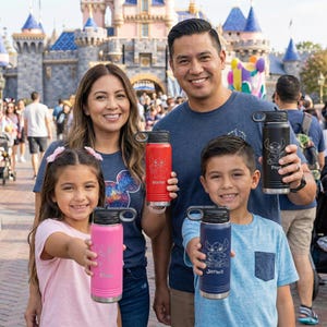 Puede incluir: Una familia posa con botellas de agua personalizadas frente a un castillo. Las botellas son de varios colores: rosa, rojo, negro y azul. Cada botella tiene una tapa negra y presenta un dise&ntilde;o de personaje y un nombre.