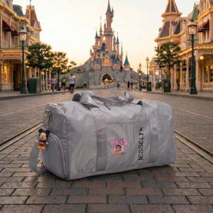 Może przedstawiać: Szara torba podr&oacute;żna z motywem Minnie Mouse i nadrukiem "JESSELYN". Torba posiada pasek na ramię i brelok z Myszka Miki. W tle zamek i ulica.
