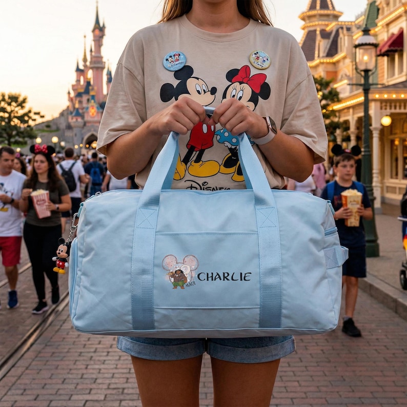 May include: A light blue duffel bag with a Mickey Mouse graphic and the name "Charlie" embroidered on the front. The bag has two handles and a shoulder strap. A Mickey Mouse keychain hangs from the zipper. The bag is held in front of a theme park.