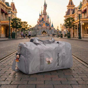 May include: A gray duffel bag with a Mickey Mouse keychain and a Disney-themed design. The bag has a side pocket and a shoulder strap. The word "JAMESON" is printed vertically on the side. The background features a castle and a street.