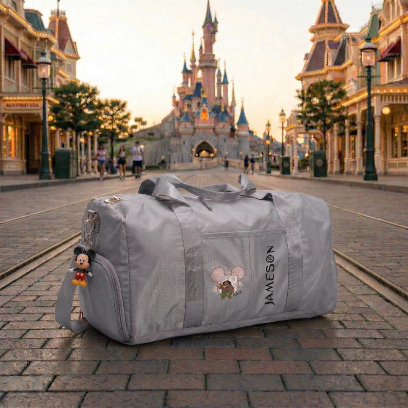 May include: A gray duffel bag with a Mickey Mouse keychain and a Disney-themed design. The bag has a side pocket and a shoulder strap. The word "JAMESON" is printed vertically on the side. The background features a castle and a street.