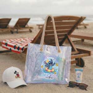 May include: A clear tote bag with rope handles featuring a Stitch graphic, a white baseball cap with a Mickey Mouse design, sunglasses, and sunscreen on a sandy beach. The bag has the text "ONLY pool vibes".