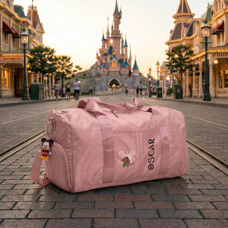 May include: A dusty rose duffel bag with a shoulder strap and a Mickey Mouse keychain. The bag has the name "OSCAR" printed on the side and a small embroidered design. The background features a castle and a street.