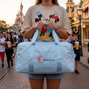 Może przedstawiać: Jasnoniebieska torba podr&oacute;żna z grafiką Minnie Mouse i nadrukiem "Angela". Torba ma dwa uchwyty i pasek na ramię. Na zdjęciu widać r&oacute;wnież osobę w beżowej koszulce z grafiką Myszki Miki i Myszki Minnie.