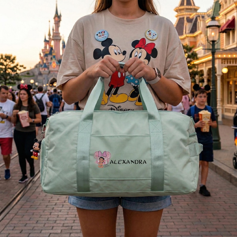 Może przedstawiać: Jasnozielona torba podr&oacute;żna z uchwytami i paskiem na ramię. Torba ma kieszeń z grafiką Minnie Mouse i nadrukowanym pod spodem imieniem "ALEXANDRA". Do torby przymocowany jest brelok do kluczy z Myszka Miki. Osoba ma na sobie koszulkę Disneya.