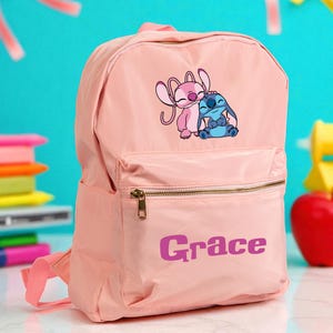 May include: A light pink backpack with a front pocket and a zippered main compartment. The backpack features a graphic of two cartoon characters and the name "Grace" in purple. The backpack has a top handle and adjustable shoulder straps.