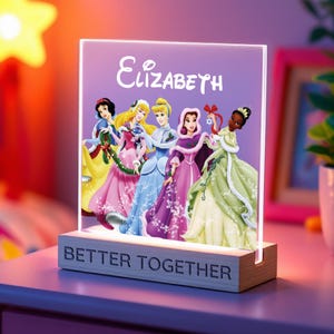 May include: A decorative night light featuring a purple panel with the name "Elizabeth" and images of animated princesses. The light sits on a wooden base with the words "BETTER TOGETHER" engraved on it.