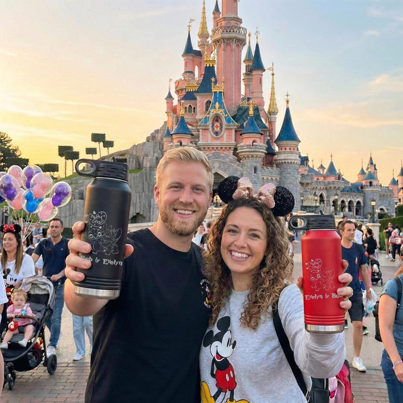 Puede incluir: Una pareja sonr&iacute;e, sosteniendo botellas de agua personalizadas frente a un castillo de Disney. Una botella es negra con un dise&ntilde;o de Stitch y la otra es roja con un dise&ntilde;o de Mickey Mouse. Las botellas tienen tapas negras.