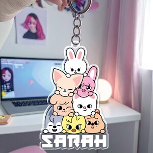 May include: A clear acrylic keychain featuring a stack of cartoon animal characters in various colors, including white, pink, yellow, and tan. The name "SARAH" is printed in white block letters at the bottom. The keychain is attached to a silver metal ring.