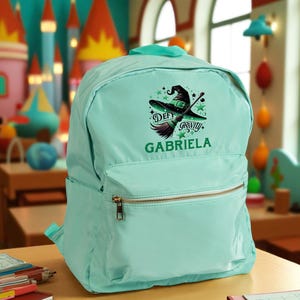 May include: A light teal backpack with a gold zipper and a top handle. The front of the backpack features a graphic of a witch's hat, broom, and the words "Defy Gravity" and "Gabriela".