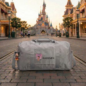 Może przedstawiać: Szara torba podr&oacute;żna z paskiem na ramię i brelokiem z Myszka Miki. Torba ma grafikę Minnie Mouse i napis "MADISON". W tle widać zamek i ulicę.