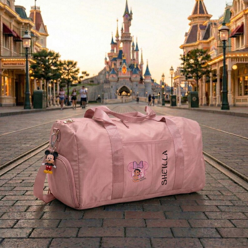 Może przedstawiać: R&oacute;żowa torba podr&oacute;żna z paskiem na ramię i brelokiem Myszki Miki. Torba ma kieszeń z grafiką Myszki Minnie i nadrukowanym imieniem "SHEILLA". Torba leży na brukowanej drodze z zamkiem w tle.