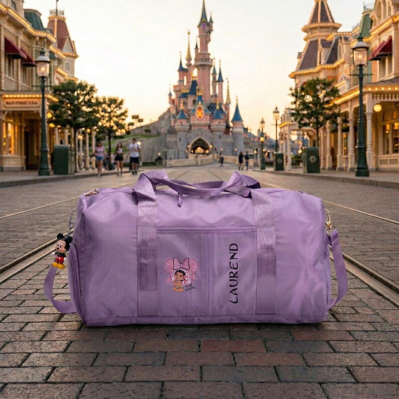 Może przedstawiać: Lawendowa torba podr&oacute;żna z paskiem na ramię i małą przednią kieszenią. Torba posiada grafikę Minnie Mouse i pionowo wydrukowane imię "LAUREND". Z boku wisi brelok z Myszka Miki. W tle widać zamek i ulicę.