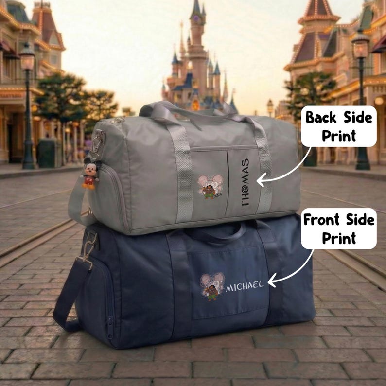 May include: Two duffel bags, one gray and one navy blue, stacked on top of each other. The gray bag has the name "Thomas" printed vertically and a cartoon graphic. The navy bag has the name "Michael" printed and a similar graphic. A Mickey Mouse keychain hangs from the gray bag.