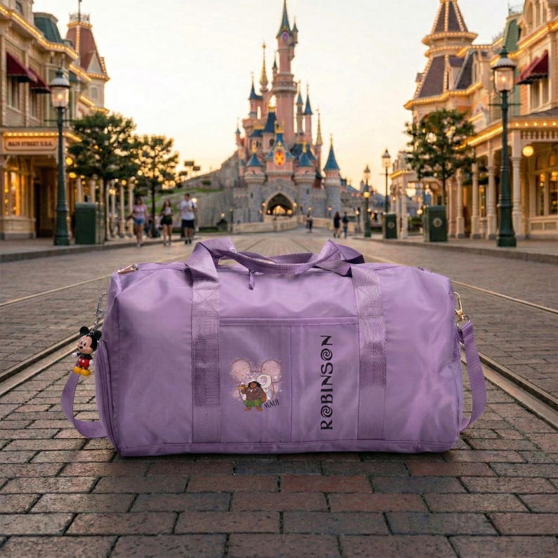 May include: A lavender duffel bag with a shoulder strap and a Mickey Mouse charm. The bag features a front pocket with a graphic of a character and the name "ROBINSON" printed vertically. The background shows a castle and a street.