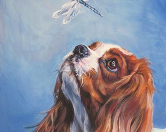 May include: A painting of a Cavalier King Charles Spaniel dog looking up at a dragonfly with blue wings. The dog has brown and white fur and is painted in a realistic style. The background is a light blue sky. The painting is signed "L.A. SHEPARD ART©".