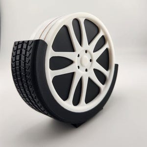 May include: A set of four white and black car wheel-shaped coasters. The coasters are stacked in a black holder, with a tire tread design on the side. The wheel design features a white rim with black spokes and a black center.