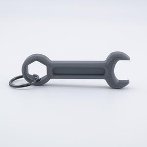 May include: A gray wrench-shaped keychain. The keychain has a hexagonal opening on one end and a curved opening on the other. A black metal ring is attached for keys. The item is made of a solid, matte material.