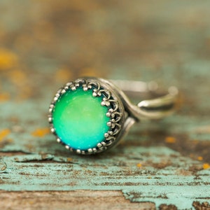Könnte beinhalten: Silberfarbener Ring mit einem großen, runden, farbwechselnden Stein. Der Stein wechselt von Grün zu Blau. Der Ring hat eine dekorative Lünettenfassung im Filigranstil. Das Ringband hat ein einzigartiges, geschwungenes Design.