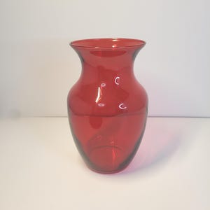 Könnte beinhalten: Eine rubinrote Glasvase mit ausgestelltem Rand und rundem Körper. Die transparente Vase steht auf einer weißen Oberfläche. Die Vase ist ein dekorativer Gegenstand, ideal für Blumen.