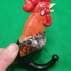 Puede incluir: Un colgador de pared con forma de gallo pintado a mano. El colgador presenta una cresta y barbilla rojas, cuerpo naranja y negro, y un gancho curvo negro. El gallo está sobre un fondo verde.