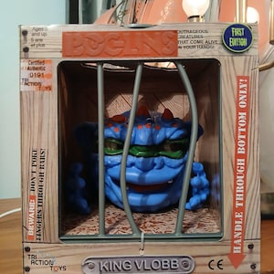 Può includere: Un giocattolo "Boglins" in scatola, "King Vlobbo", un mostro blu con occhi verdi e corna rosse, è in gabbia. La scatola recita "First Edition" e "Outrageous Creatures That Come Alive In Your Hands!" con avvertenze e il logo Tri Action Toys.