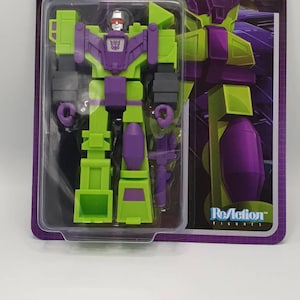 May include: A retro-style Devastator action figure from the Transformers series, predominantly green and purple. The packaging features the Transformers logo and the phrase "MORE THAN MEETS THE EYE!" The figure is in a clear plastic case.