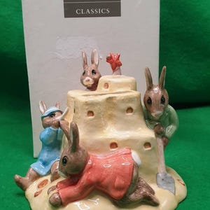 Puede incluir: Figurita de Royal Doulton Classics con conejos alrededor de un castillo de arena. Un conejo lleva un abrigo rojo, otro un vestido azul y un tercero está cavando con una pala. El castillo de arena tiene una estrella roja encima.