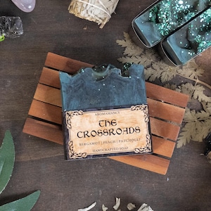 Può includere: Una saponetta artigianale di colore verde acqua scuro con un'etichetta decorativa che recita "The Crossroads". Il sapone è su un portasapone di legno. L'etichetta elenca i profumi: bergamotto, pesca e patchouli. Lo sfondo include cristalli ed erbe essiccate.