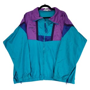 May include: A vintage Columbia windbreaker jacket in teal, purple, and navy blue. The jacket has a full zipper, a high collar, and the Columbia logo on the chest. The color-blocked design features teal sleeves and body, with purple and navy accents.