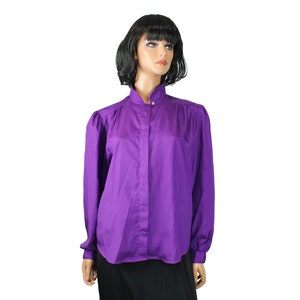 Op de afbeelding: Een paarse blouse met lange mouwen, een hoge opstaande kraag met één zichtbare knoopsluiting, een verborgen knoopsluiting aan de voorzijde en geplooide details bij de schouders, gepresenteerd op een etalagepop met een zwarte rok.