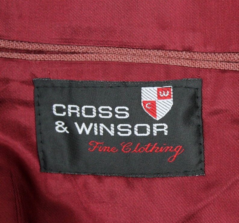 May include: A black label with white text that reads "CROSS & WINSOR Fine Clothing". A red and white crest with a "C" and a "W" is above the text.