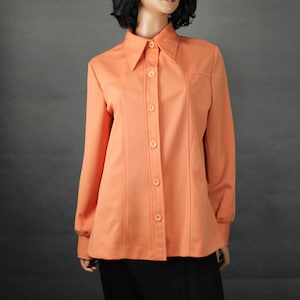 May include: A peach-colored button-down shirt with a pointed collar and long sleeves. The shirt has vertical seams down the front and is paired with a black skirt. The mannequin is wearing a black wig.