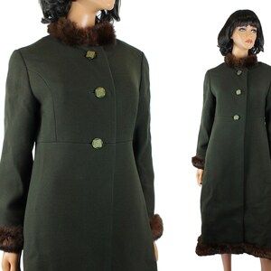 May include: A dark olive green long coat featuring a brown faux fur collar and matching faux fur trim at the cuffs and hem. The coat has a high neckline, long sleeves, and a front button closure with three decorative green buttons.