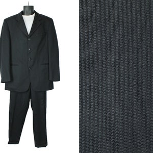 Può includere: Un abito nero con giacca monopetto e pantaloni abbinati. La giacca ha un revers a punta e tre bottoni. Il tessuto ha una texture leggermente a coste.
