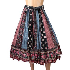 May include: A midi-length skirt featuring vertical panels of black, blue, and dusty rose fabric with various floral patterns. The skirt has a gathered waist with a matching tie belt and a wide, ruffled hemline decorated with a detailed floral border.