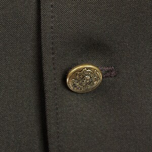 Op de afbeelding: Een donkerbruin stoffen jack met een gouden knoop met een decoratief ontwerp.