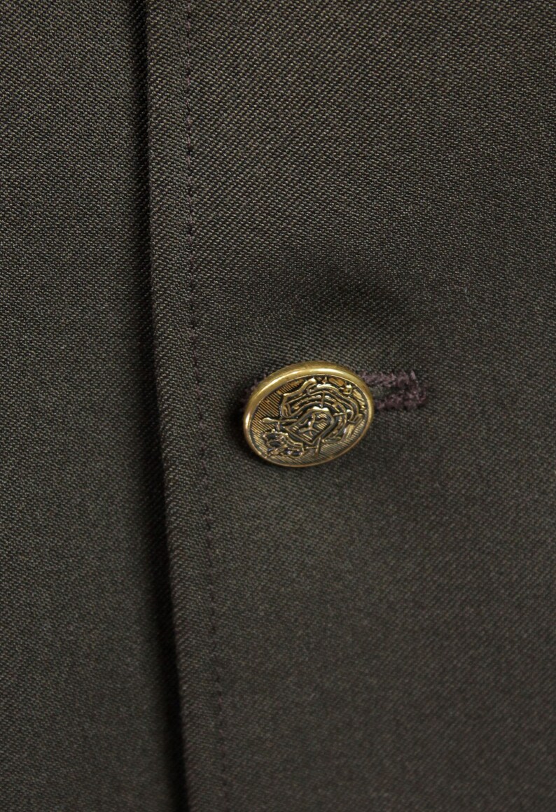 Op de afbeelding: Een donkerbruin stoffen jack met een gouden knoop met een decoratief ontwerp.