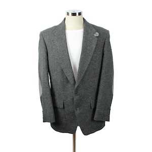 Puede incluir: Un blazer gris con coderas y una camisa blanca debajo. El blazer tiene dos bolsillos delanteros y cierre de botones. Un pequeño pin gris está en la solapa. El blazer se exhibe en un maniquí.