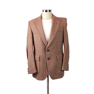 Può includere: Un blazer in tweed marrone con toppe in pelle sui gomiti e tre bottoni marroni. Il blazer ha una tasca sul petto e due tasche anteriori.