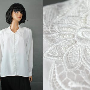 Puede incluir: Una blusa blanca con mangas largas y un delicado escote bordado. La blusa presenta pequeños botones nacarados en la parte delantera. El escote está adornado con encaje intrincado y pequeñas cuentas redondas. La prenda se exhibe en un maniquí.