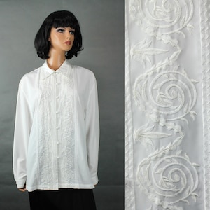 May include: White long-sleeve shirt with intricate embroidered detailing down the front and on the collar. The shirt features a button-down design and is displayed on a mannequin. The embroidery has a floral pattern.