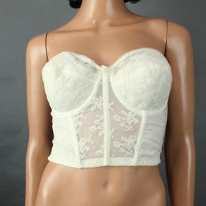 May include: Ivory-colored strapless bustier with delicate lace detailing. The bustier features a structured design with a central panel and underwire support. The lace overlay adds a touch of elegance to this lingerie piece.