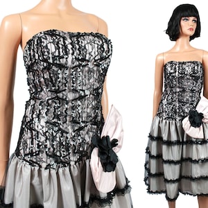 Op de afbeelding: Een strapless zwart-witte kanten jurk met een geruite rok en een roze satijnen strik. De jurk is gemaakt van een transparante stof met een zwarte kanten overlay. De rok is gelaagd en heeft een geruite zoom. De strik is aan de jurk bevestigd op de taille.