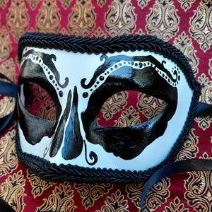 May include: A white masquerade mask featuring bold black painted designs around the eye openings and a central nose detail. The edges of the mask are trimmed with a thick black braided cord, and black satin ribbons are attached to the sides.