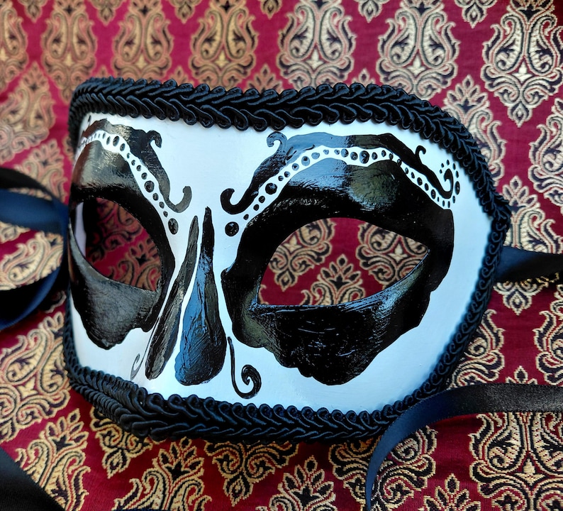May include: A white masquerade mask featuring bold black painted designs around the eye openings and a central nose detail. The edges of the mask are trimmed with a thick black braided cord, and black satin ribbons are attached to the sides.