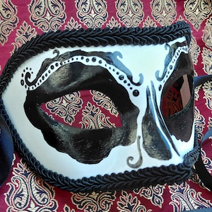 May include: A white masquerade mask featuring bold black painted accents around the eye openings and intricate black scrollwork details. The edges of the mask are trimmed with a thick black braided cord, and black satin ribbons are attached.