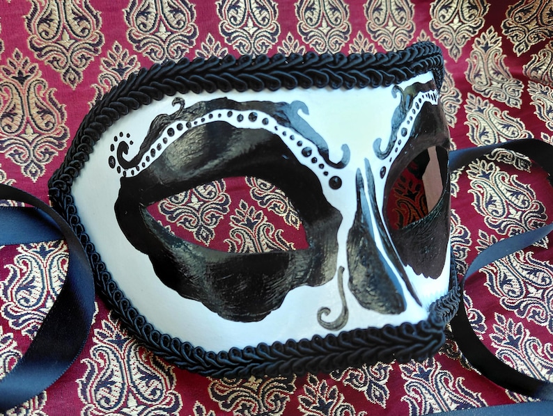 May include: A white masquerade mask featuring bold black painted accents around the eye openings and intricate black scrollwork details. The edges of the mask are trimmed with a thick black braided cord, and black satin ribbons are attached.